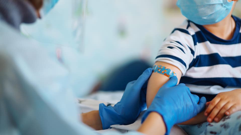 Un médecin appose un diachylon sur le bras d'un enfant. Les deux portent un masque chirurgical. 