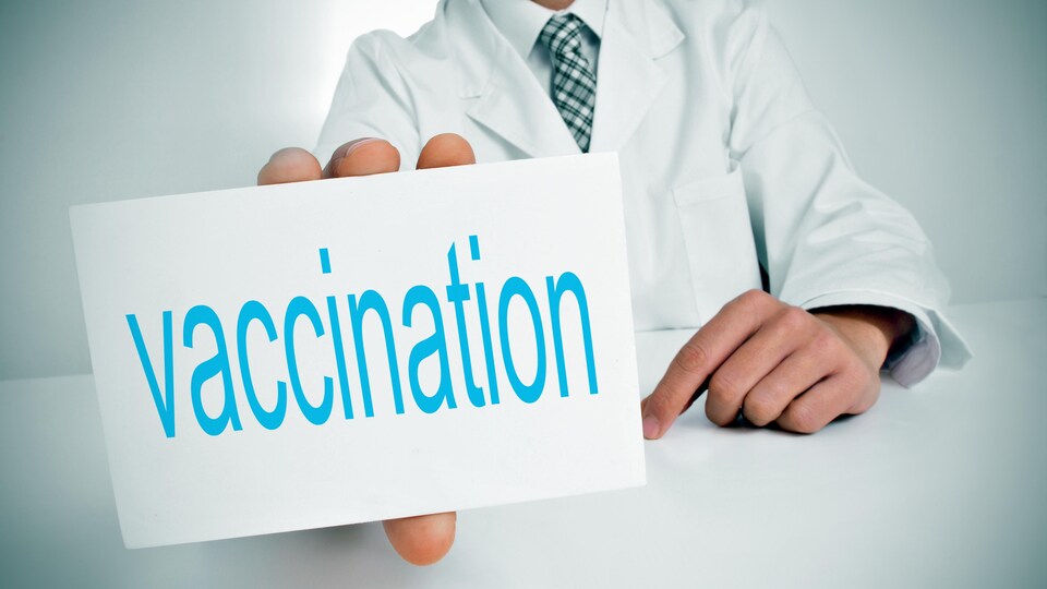 Un homme dont on ne voit pas le visage, vêtu d'une blouse blanche, tient un carton dans sa main droite où est écrit en bleu le mot vaccination.
