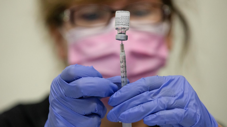 Une infirmière portant un masque rose et des gants bleus prépare une dose de vaccin.