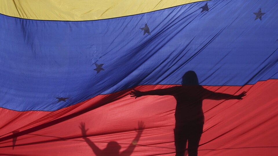 La silhouette d'une femme, debout, les bras étendus, se profile derrière le drapeau du Venezuela.