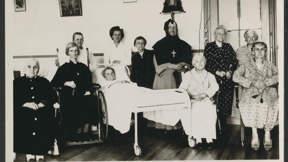 Photo en noir et blanc montrant plusieurs femmes âgées, dont une alitée et une en fauteuil roulant, en compagnie d'une religieuse.