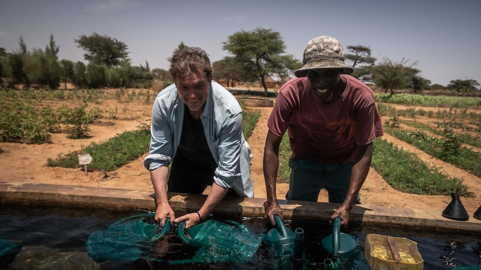 Photo de l'animateur Vincent Graton avec un agriculteur sénégalais en train de puiser de l'eau dans une oasis, souriant à la caméra.