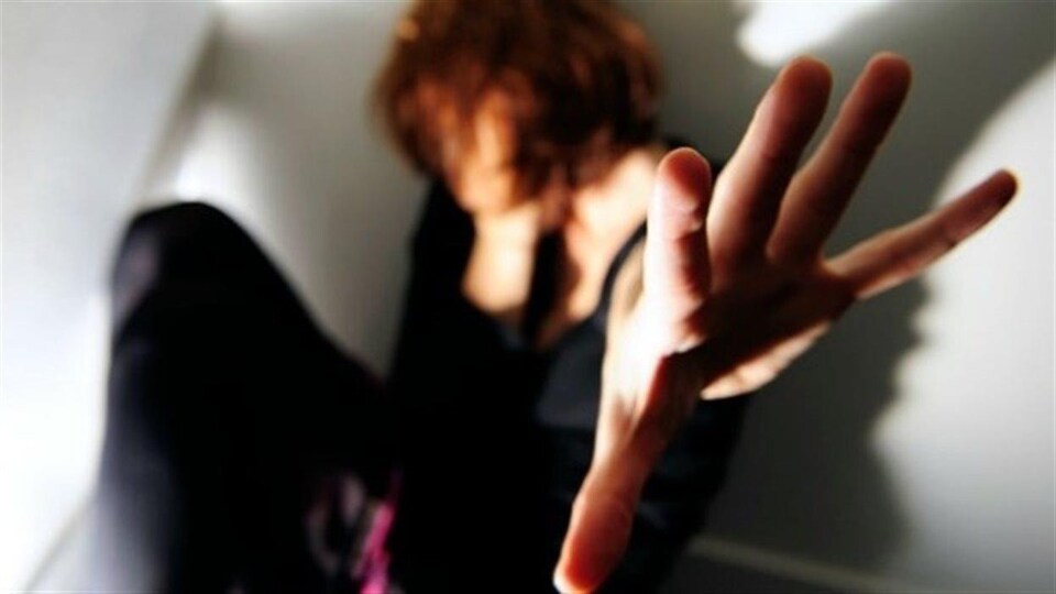 Une femme accroupie dans un coin d'une pièce, levant la main pour se protéger.