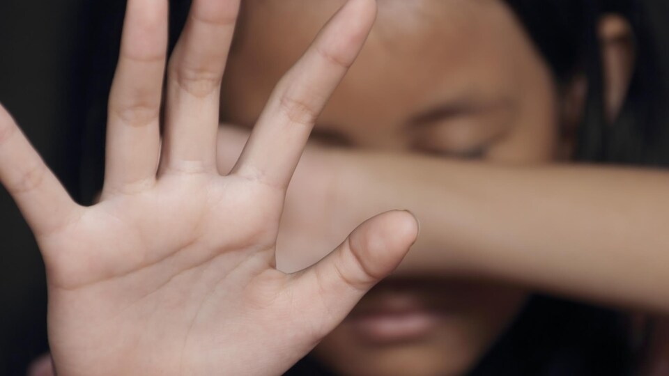 Une jeune fille se protège le visage à l'aide de son bras et fait "stop" à l'aide de son autre main.