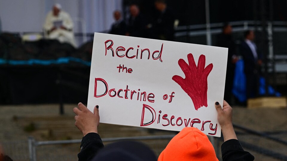 Gros plan sur une personne qui tient une pancarte de protestation sur laquelle il est écrit en anglais «Abroger la doctrine de la découverte» pendant que le pape François parle sur scène à l'école élémentaire Nakasuk, à Iqaluit, le 29 juillet 2022.