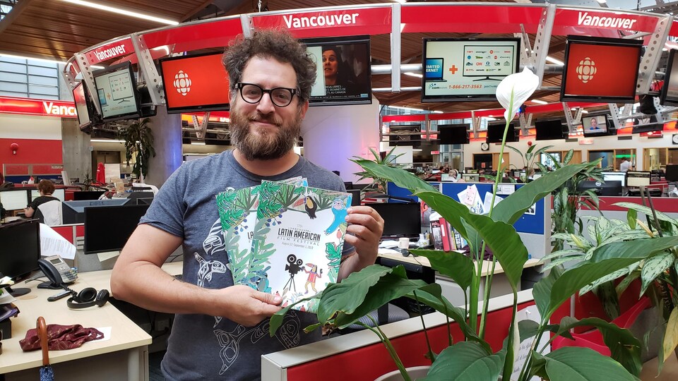 Homme à lunettes debout, près d'une plante fleurie, tenant des brochures dans un studio de télévision.