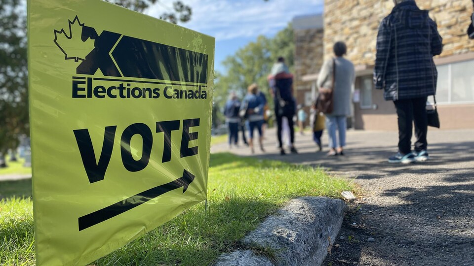 Une affiche indiquant la direction d'un bureau de vote en avant-plan et une file d'attente en arrière-plan.