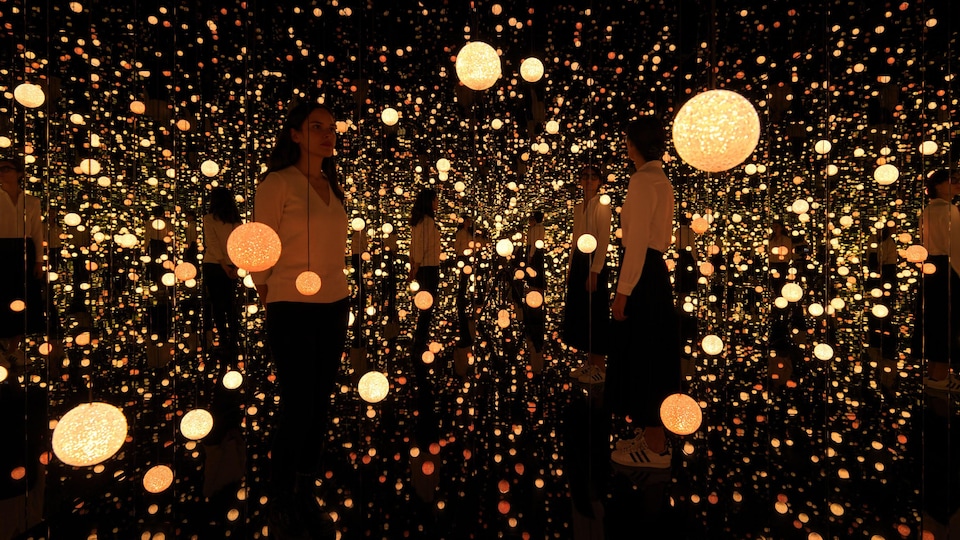 Deux personnes se tiennent dans une salle dont les murs sont des miroirs. De très nombreuses ampoules suspendues émettent de la lumière et les reflets des personnes et des objets sont infinis. 