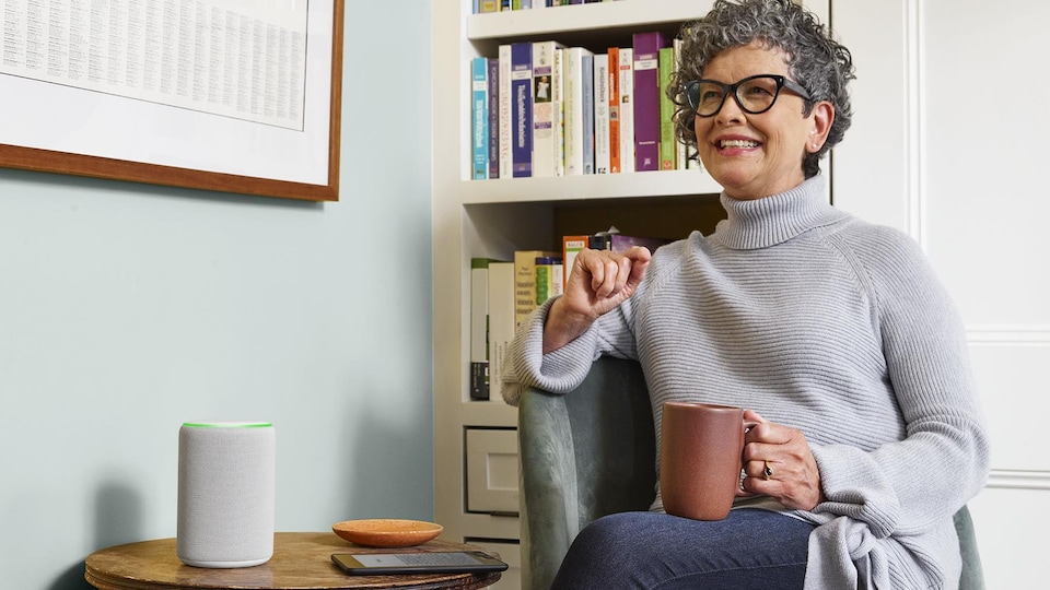 Une femme assise dans un fauteuil écoute quelque chose diffusé sur le haut-parleur Amazon Echo déposé sur une table.
