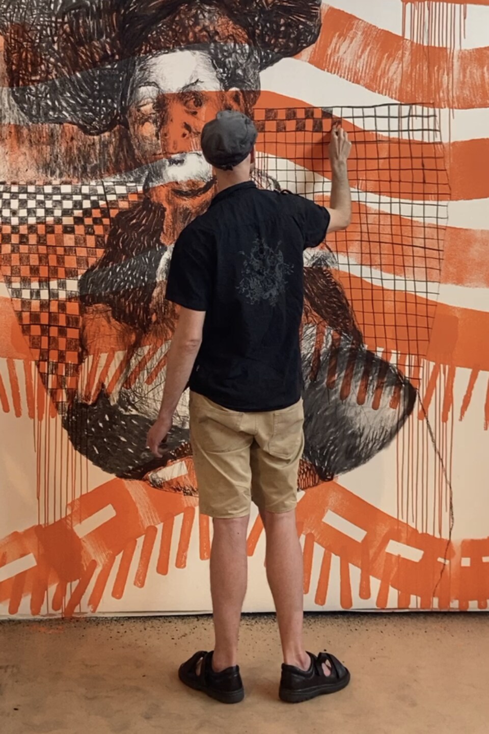 Un homme vêtu d'une casquette, d'une chemise sombre et d'un bermuda dessine sur un mur.