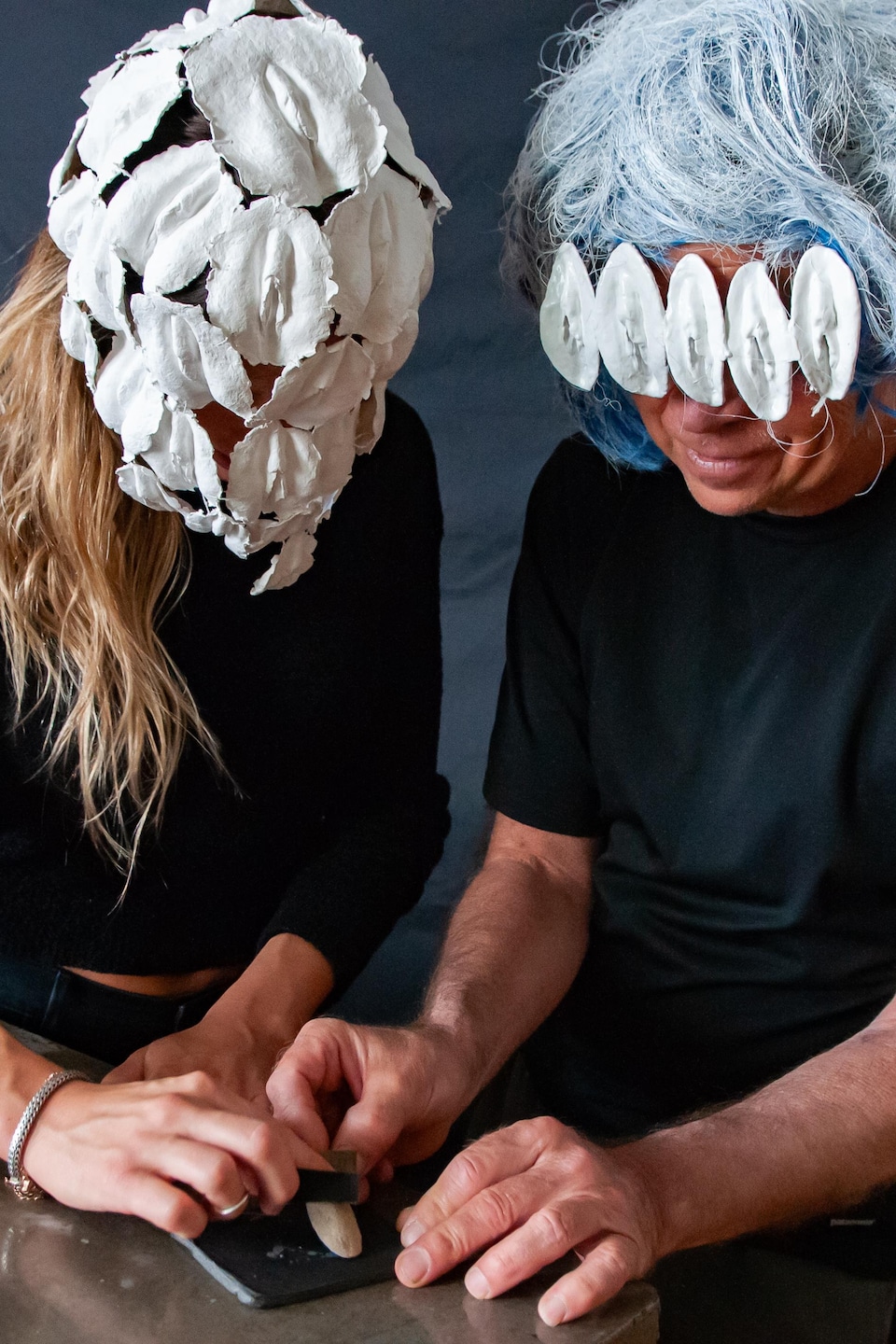 Un homme et une femme portant tous les deux des masques formés de vulves sablent une petite vulve dans une cuisine. 