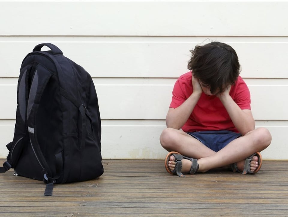 Un enfant la tête dans les mains, assis par terre près de son sac d'école.