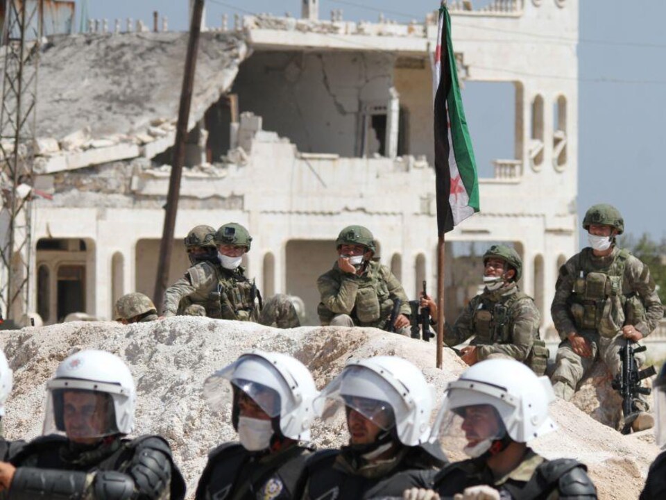 Deux rangées de soldats avec des masques. À l'arrière-plan, un drapeau syrien et un bâtiment à moitié démoli. 