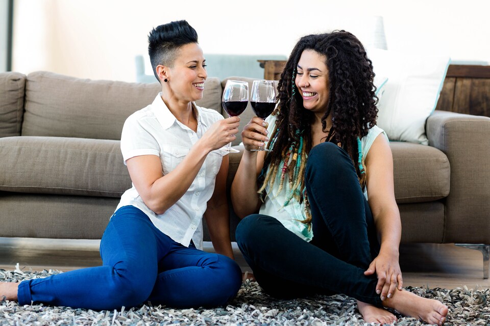 Deux femmes souriantes, des coupes de vin rouge à la main, assises par terre sur un tapis près d'un sofa. 