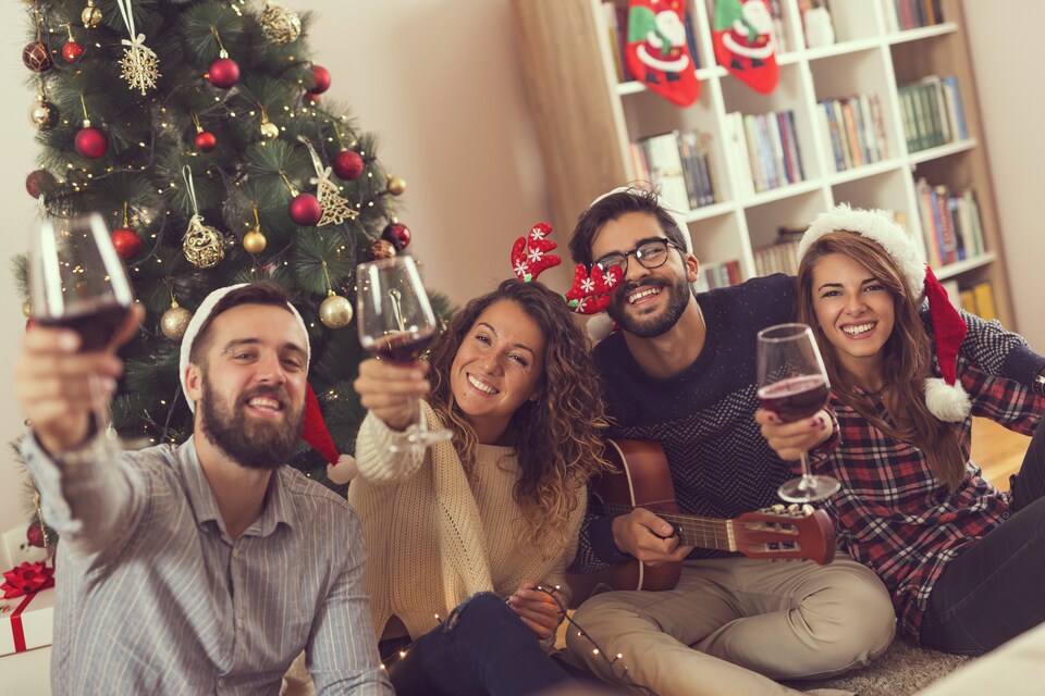Un groupe de 4 personnes dans un salon décoré pour Noël prennent un verre de vin rouge en souriant. 