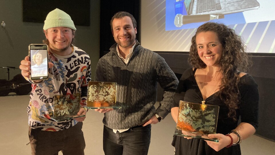 Pierre-Philippe Côté, Nicolas Zemmour et Ariane Deslions tiennent des trophées. Pierre-Philippe tient aussi son téléphone, où l'on voit une photo de Valérie Whissel.
