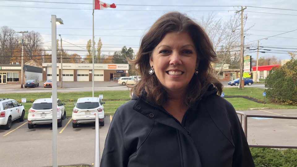 Une femme souriante portant un manteau noir, devant l’édifice de Radio-Canada, à Charlottetown. Quelques voitures stationnées et le drapeau du Canada se trouvent au fond.