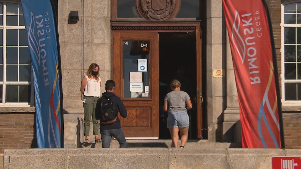 La porte d'entrée du Cégep de Rimouski à côté de laquelle se tient une femme qui regardent deux étudiants monter les marches pour entrer dans l'établissement.