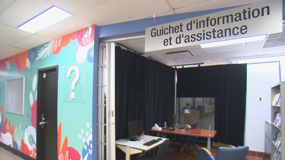 Une femme attend à un bureau. Il est écrit « guichet d'information et d'assistance » sur une affiche au-dessus de son bureau.