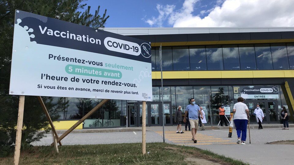 Des gens masqués marchent devant le Centre de foires. Une affiche indique "Vaccination COVID-19 : présentez-vous seulement 5 minutes avant l'heure de votre rendez-vous".