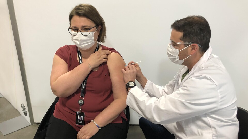 Un homme vêtu d'un sarrau blanc et portant un masque de procédure blanc ainsi que des lunettes de protection est assis à la gauche d'une dame et la vaccine. Celle-ci porte un chandail rouge, un masque de procédure blanc et des lunettes. Elle semble concentrée, comme elle se fait vacciner.