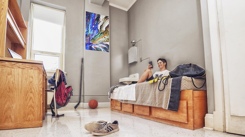 Une chambre d'étudiants avec des souliers sur le plancher, un ballon sur le sol, un bâton de hockey sur le mur et un étudiant dans son lit, sur son téléphone.