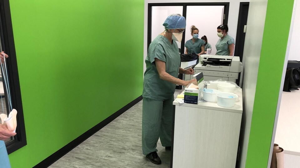 Trois professionnels de la santé portant le masque de procédure et un uniforme médical oeuvrent à la clinique de dépisage de Plessisville, dont les mûrs sont d'un vert éclatant. L'un des employés travaille à une table où sont posés une boîte de masques ainsi que plusieurs récipients blancs dans lesquels se trouvent des instruments de travail.