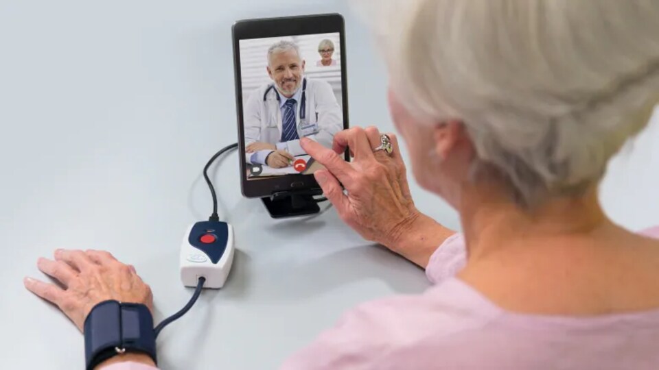 Une patiente parle à un médecin à travers une tablette numérique. Son bras est connecté à un appareil d'auscultation.