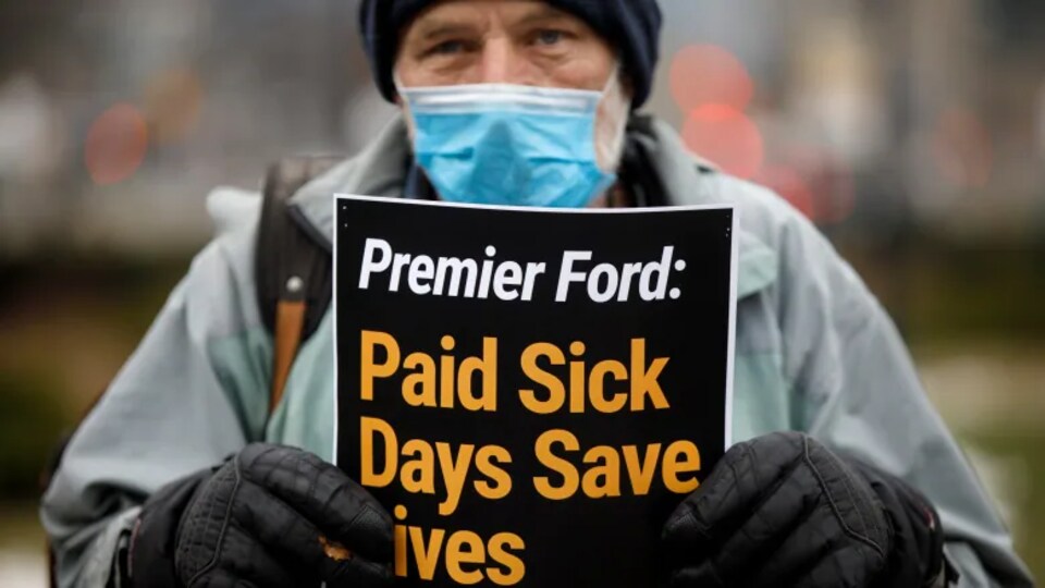 Un manifestant en tenue d'hiver tient une pancarte demandant au premier ministre Ford de mettre en place des congés de maladie payés.