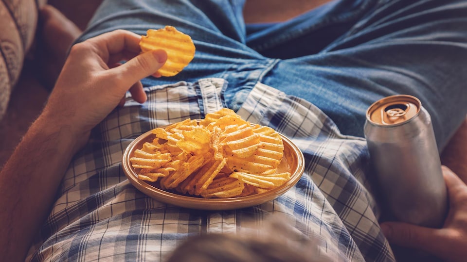 Un homme couché sur un divan en train de manger des croustilles qui sont dans une assiette sur son ventre. Il tient une canette dans sa main droite.
