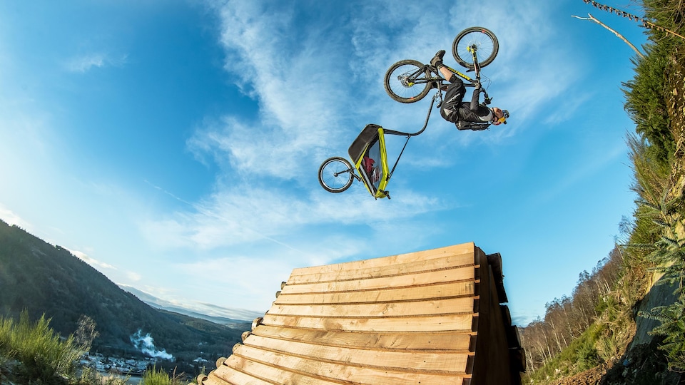 Image tirée du film Danny Daycare, du cycliste et photographe Danny MacAskill. On y voit un cycliste dans les airs, en train de faire un tour sur lui-même, avec un chariot pour enfant attaché à son vélo.