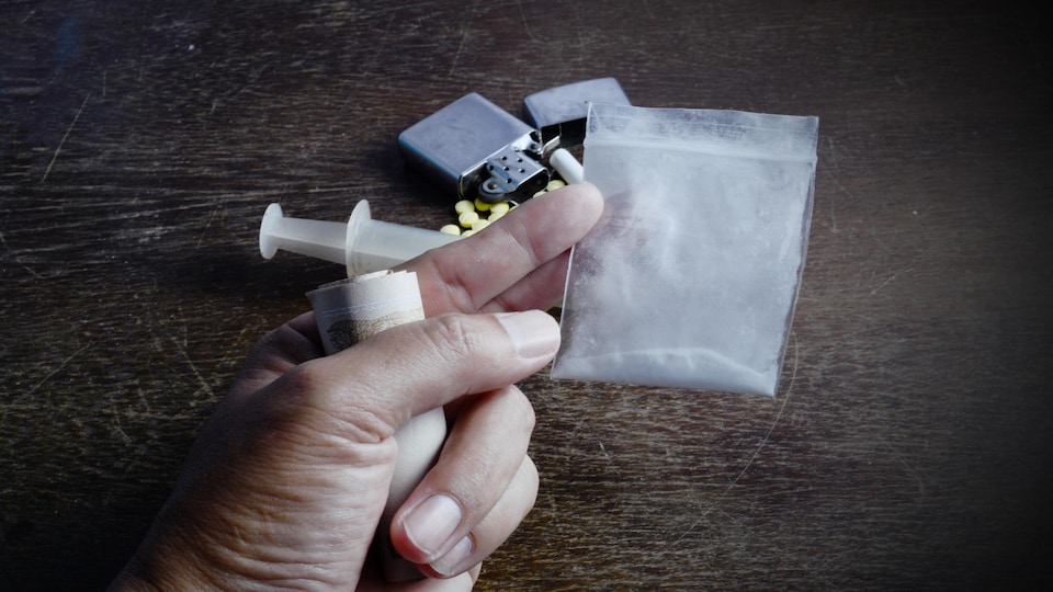 Une main qui tient de l'argent et de la poudre d'héroïne dans un sac. Une seringue, un médicament, une cigarette et un briquet sont déposés sur une table à l'arrière-plan.