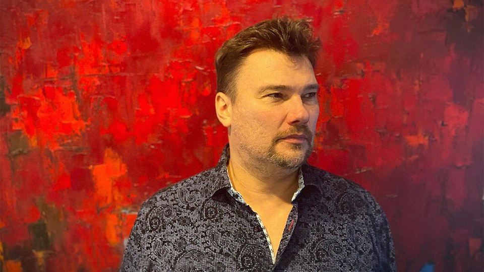 L'artiste Dominik Sokolowski qui regarde vers la droite devant une de ses toiles.