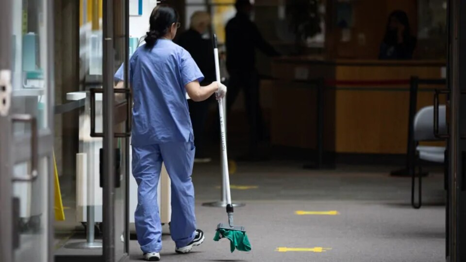 Une femme en tenue médicale déambule dans un bâtiment avec un balai en main.