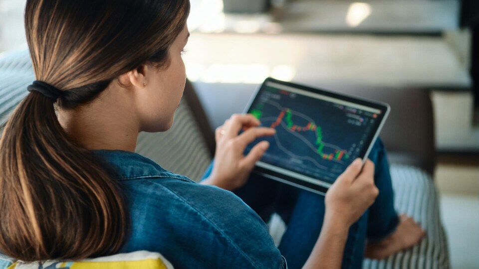 Une jeune femme assise sur un fauteuil regarde une tablette qui démontre des informations de finances.