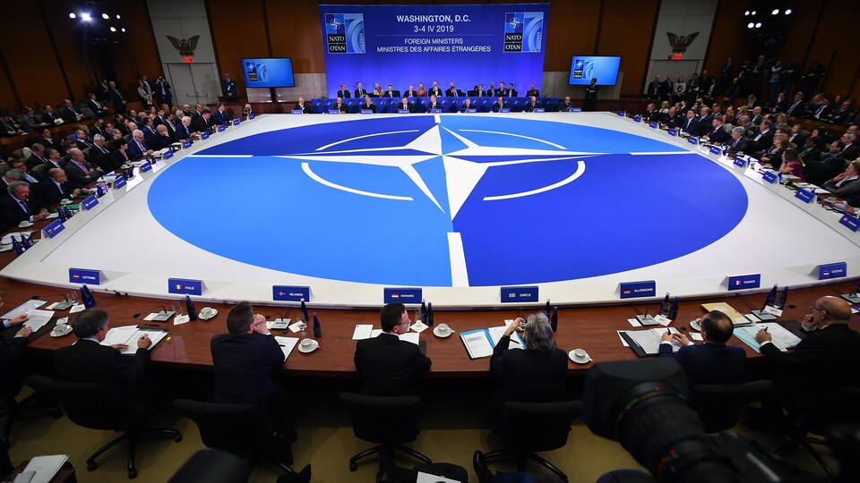 Des représentants des membres de l'OTAN assis à une énorme table sur laquelle le logo de l'organisation est à l'honneur.