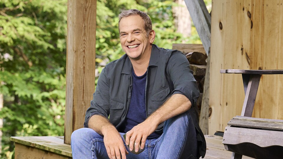 Garou, souriant, assis sur un balcon devant une forêt.