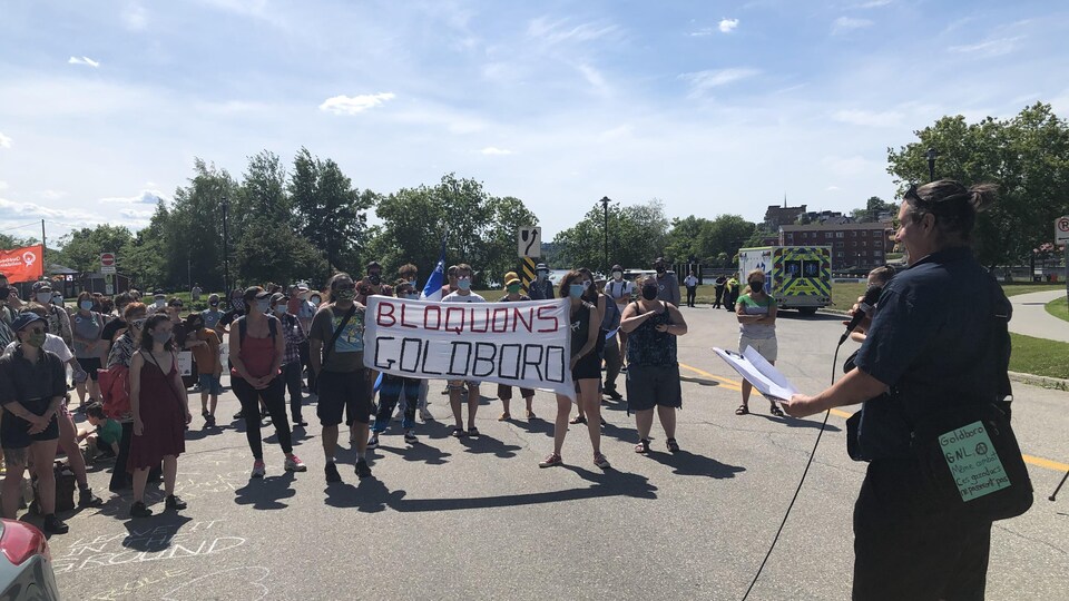 Un homme parle dans un micro, s'adressant à une foule de militants portant un drap avec l'inscription « Bloquons Goldboro »