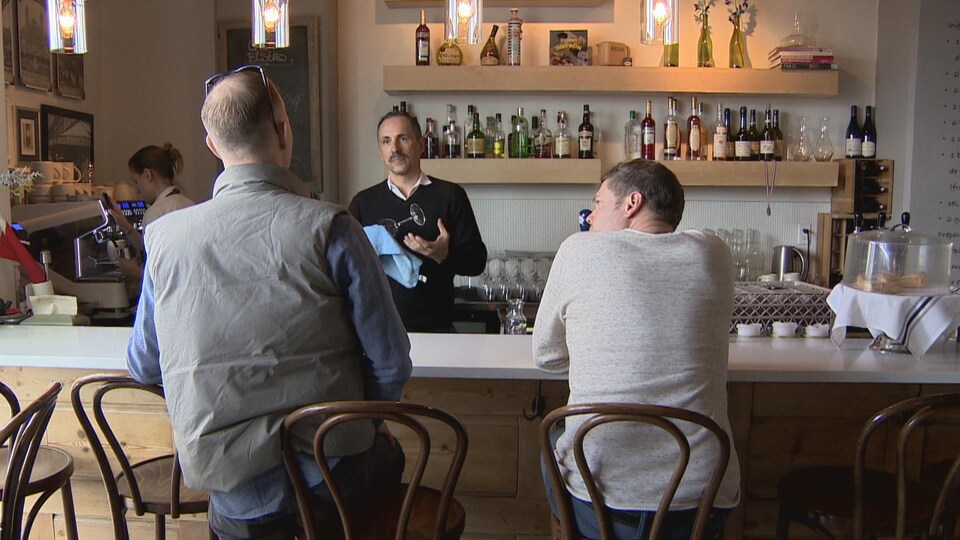Un homme parle à deux clients d'un restaurant à un bar. Les deux clients sont de dos. L'homme, Gilles Brassard, astique un verre qu'il tient dans sa main de l'autre côté du bar.