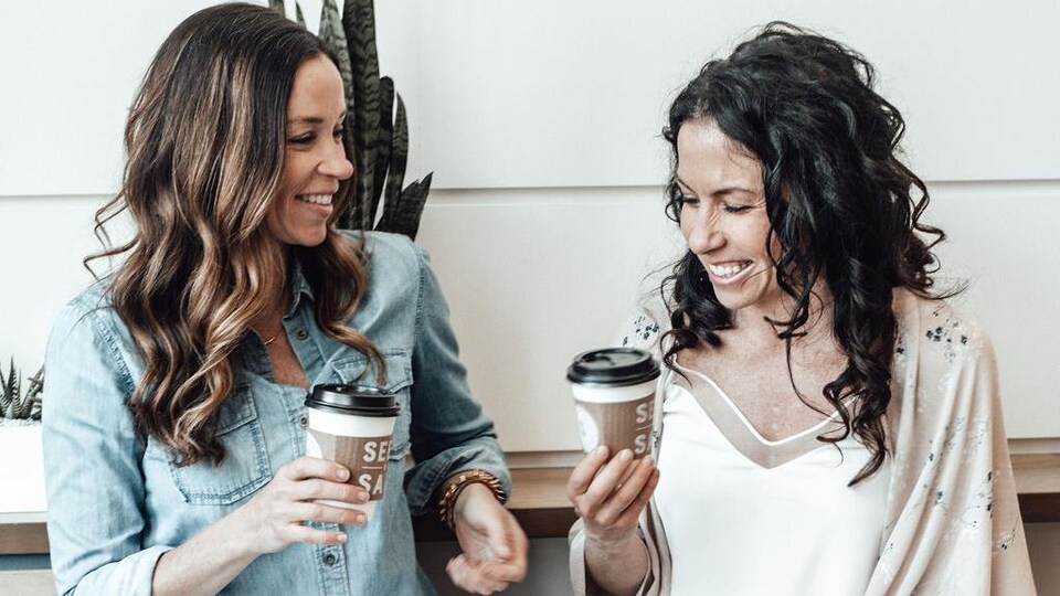 The two women are seated on stools holding cups of coffee stamped with their restaurant's brand. 