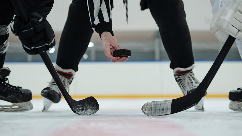 Un arbitre tenant une rondelle de hockey au-dessus d'une patinoire avec deux joueurs qui tendent leurs bâtons.