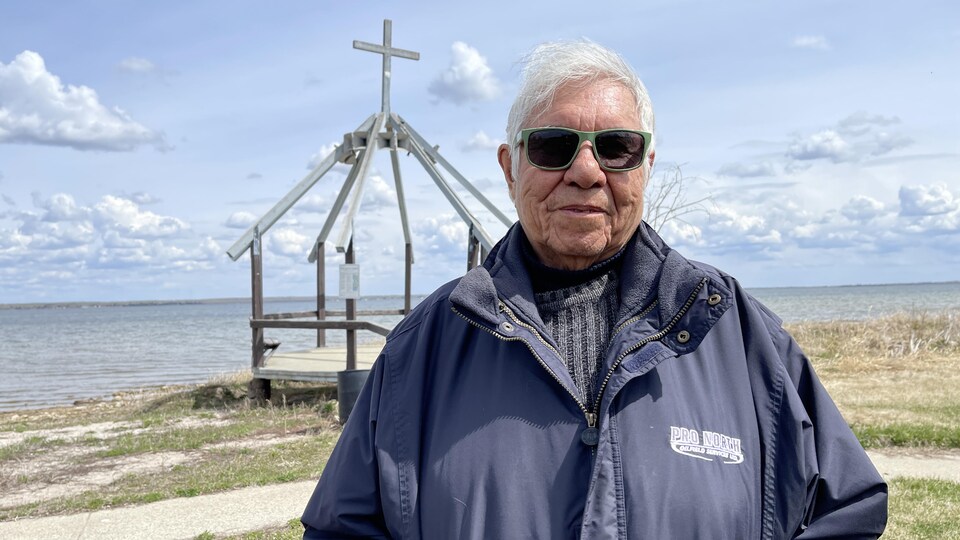 Jack Lloyd Wepataham pose au bord du lac devant une structure de bois surmontée d'une croix. 