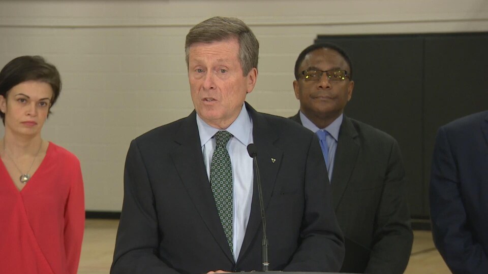 Le maire de Toronto John Tory parle devant un micro dans un gymnase. En second plan, des intervenants attendent de prendre la parole.