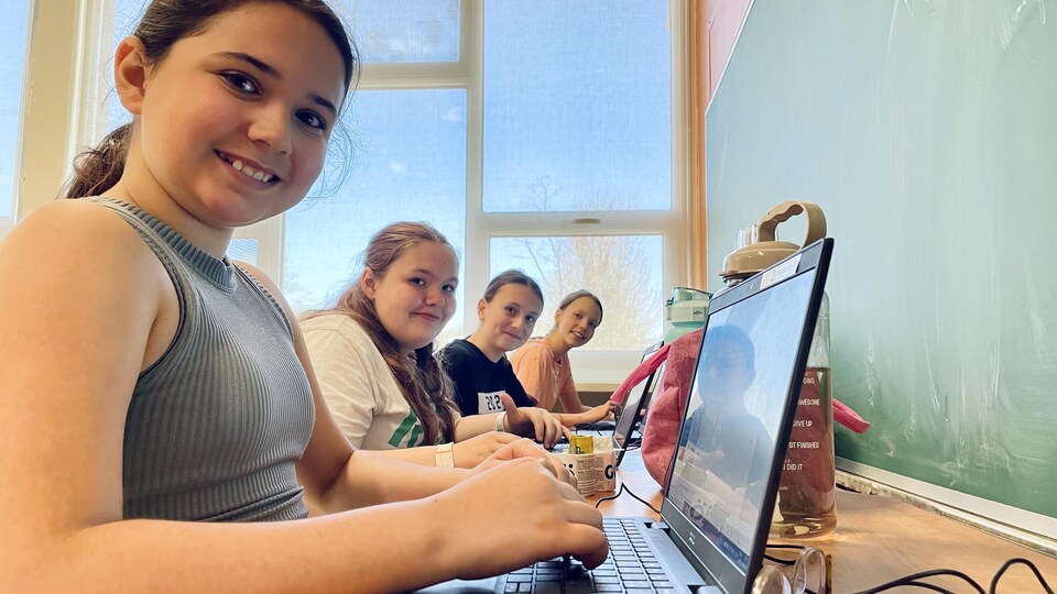 Quatre pré-adolescentes sourient, attablées devant des ordinateurs portables.