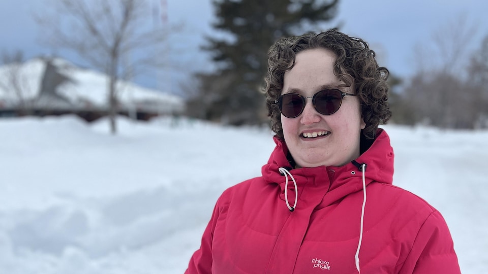 Julie Bourque face à la caméra, en manteau d'hiver et lunette de soleil, avec de la neige en arriière-plan.