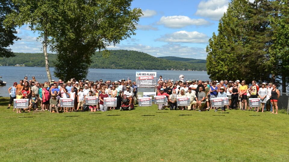 Plusieurs dizaines de personnes tenant des affiches « Lac-des-Plages incompatible à l'activité minière » devant un plan d'eau.