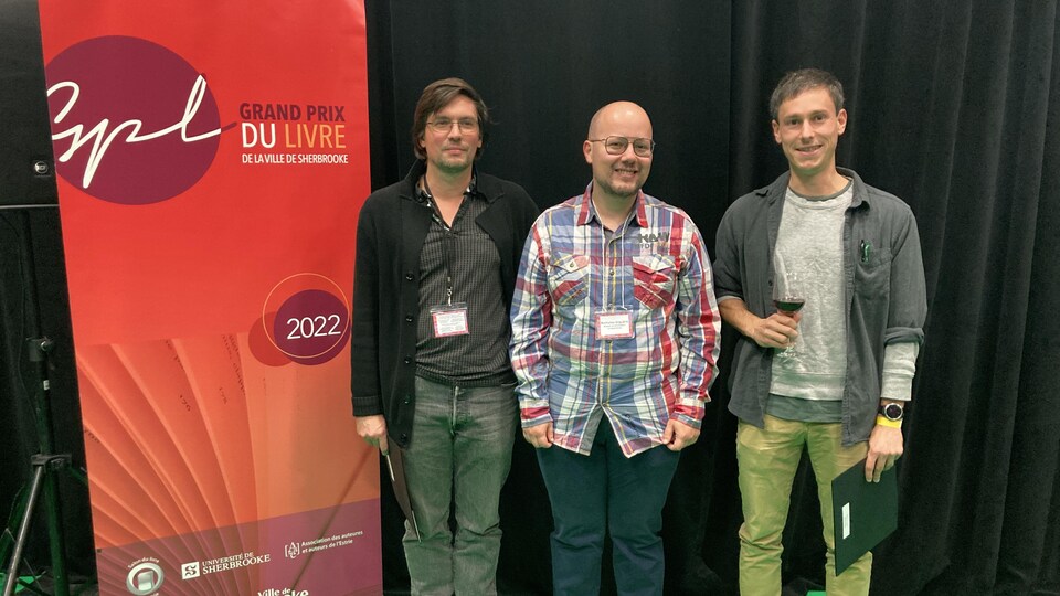 Les trois lauréats sourient à côté d'une affiche du grand prix. 