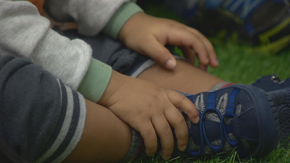 Des mains et un pied d'un jeune enfant à la peau foncée. L'enfant porte des vêtements de coton ouaté et des chaussures de sport neuves. Son chandail comporte une minuscule tache rougeâtre sur la manche. Il est assit dans l'herbe.