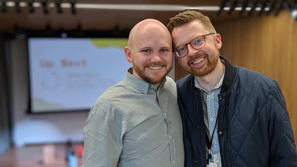 Deux hommes se tiennent côte à côte et sourient à la caméra. L'un est chauve et porte une chemise. L'autre a des lunettes et porte un blouson.