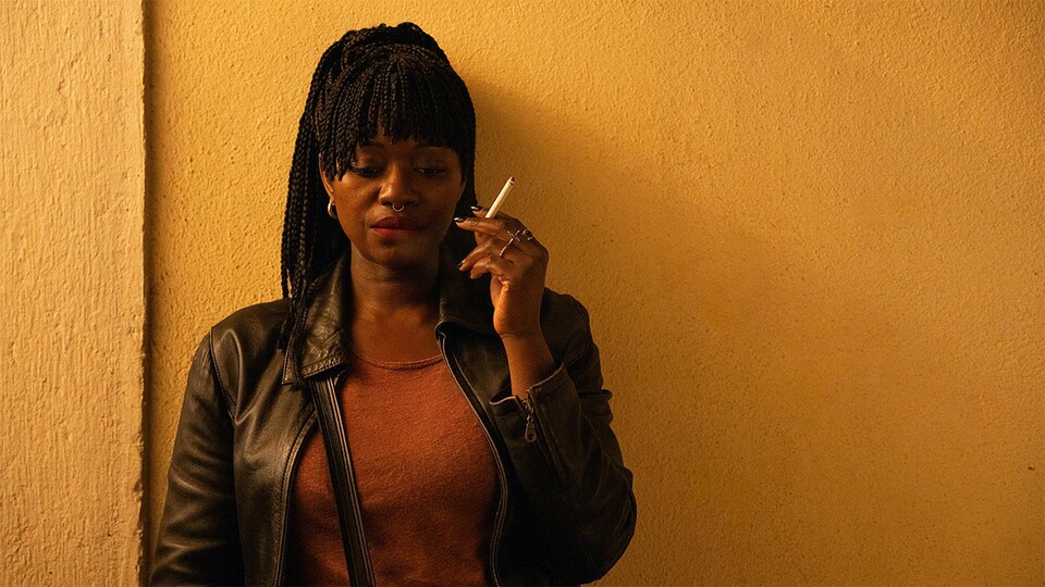 Une femme appuyée sur un mur qui tient une cigarette dans sa main gauche.